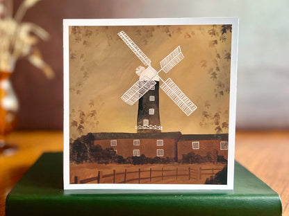 Premium illustrated greeting card featuring Skidby Windmill in East Yorkshire behind farm buildings under an autumnal orange sunset sky, displayed standing upright on a tabletop. Countryside landscape art greeting card inspired by rural Yorkshire scenery.
