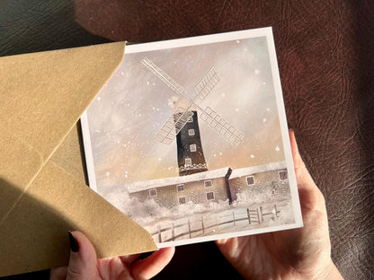 Premium illustrated greeting card featuring Skidby Windmill in East Yorkshire standing behind snow-covered farm buildings under a winter sunset sky with pink and purple tones. Snow is falling across the scene, creating a festive countryside Christmas card design inspired by rural Yorkshire.