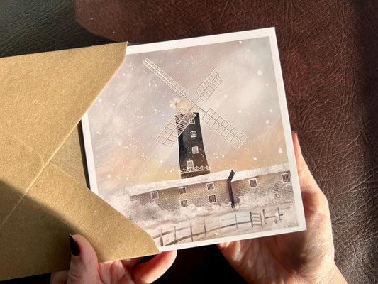 Premium illustrated greeting card featuring Skidby Windmill in East Yorkshire standing behind snow-covered farm buildings under a winter sunset sky with pink and purple tones. Snow is falling across the scene, creating a festive countryside Christmas card design inspired by rural Yorkshire.
