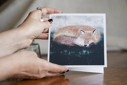 Premium illustrated art greeting card featuring a sleeping fox in a calm woodland scene, set against a textured dark green and grey background, held in hands facing the camera to show the full front design. Versatile wildlife art card suitable for any occasion.