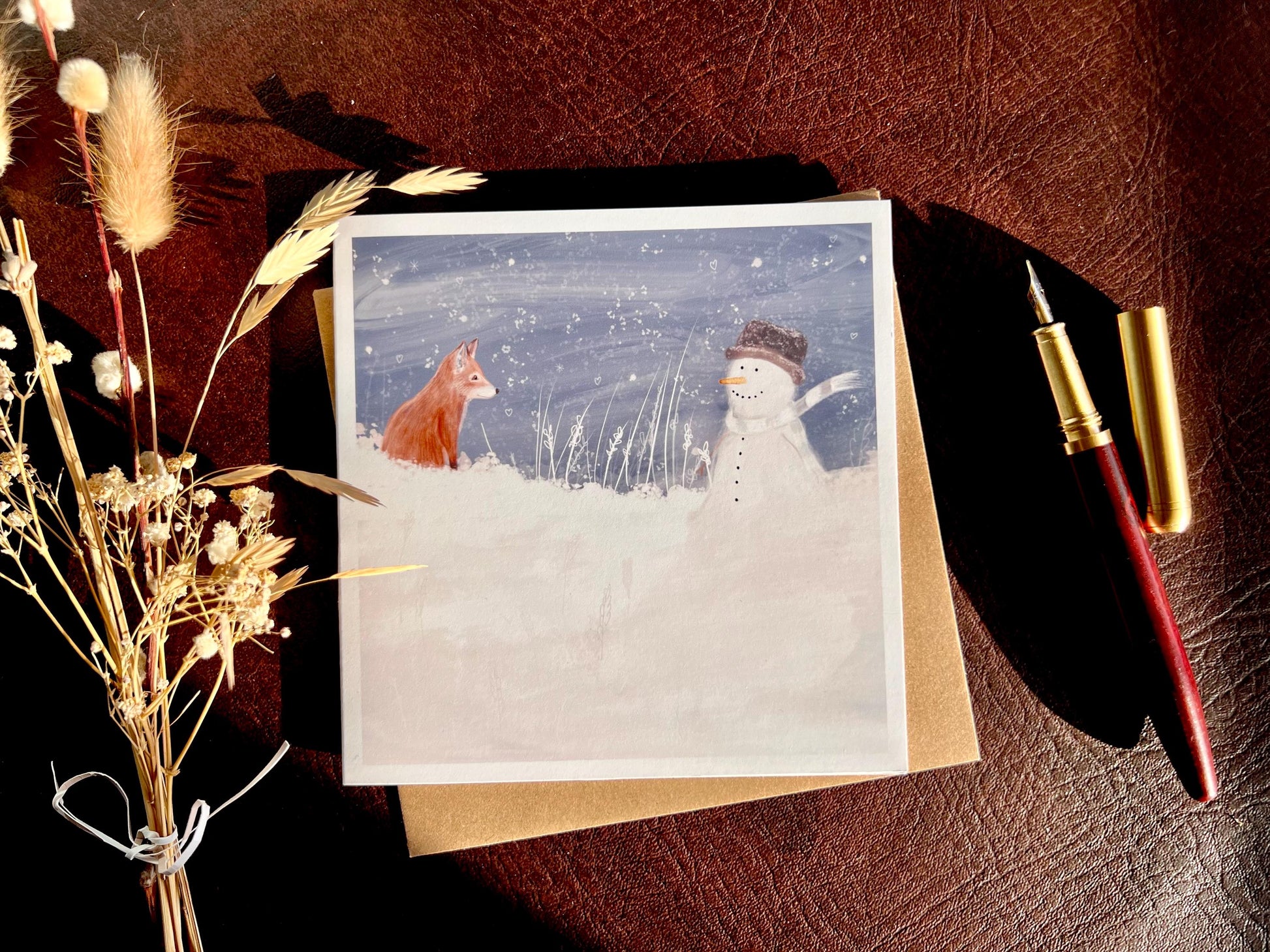 Premium illustrated Christmas greeting card featuring a happy snowman wearing a hat looking at a fox cub in a snowy winter landscape, laid flat on a leatherette tabletop with a recycled fleck kraft envelope and pen. Festive wildlife-inspired seasonal greeting card flat lay.