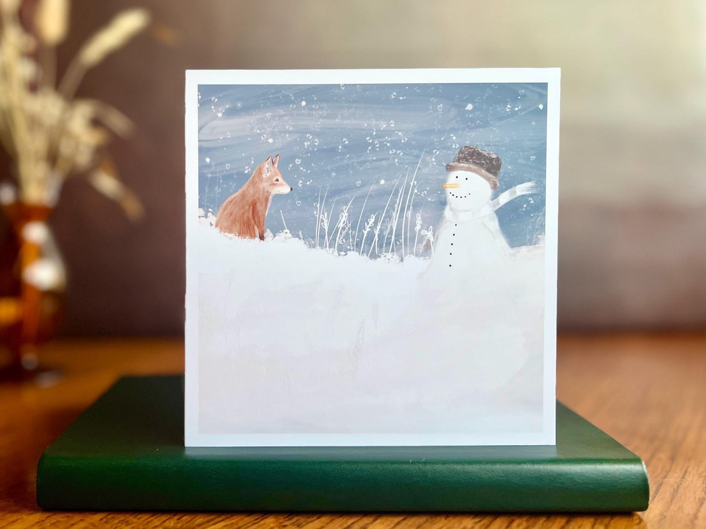 Premium illustrated Christmas greeting card featuring a happy snowman wearing a hat looking at a fox cub in a snowy winter landscape, displayed standing on a green leatherette book placed on a wooden tabletop. Dried flowers appear in the background, creating a styled festive seasonal scene.