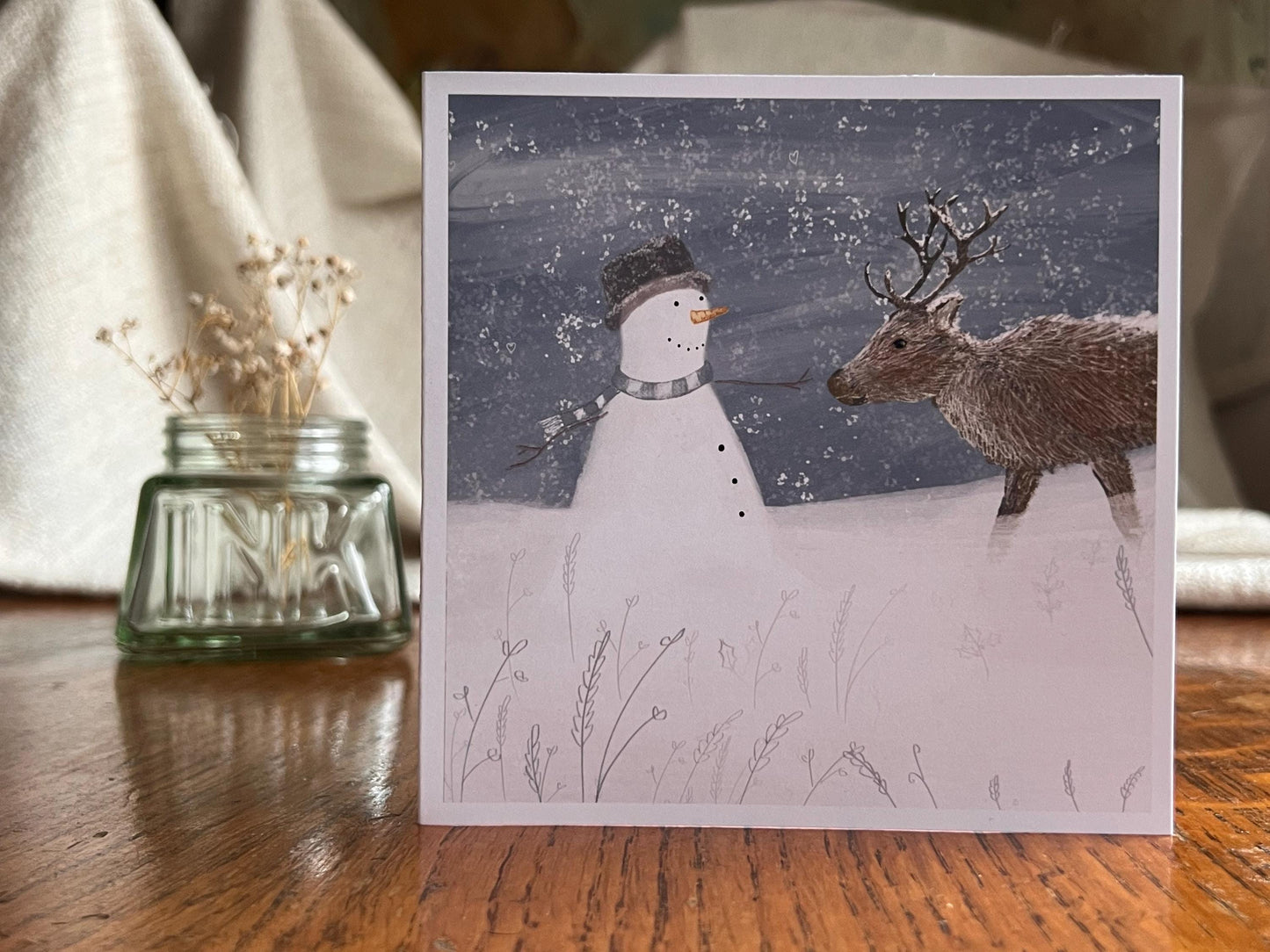 Premium illustrated Christmas greeting card featuring a happy snowman wearing a hat looking at a reindeer in a snowy winter landscape, displayed standing upright on a tabletop. Festive wildlife-inspired seasonal greeting card design.