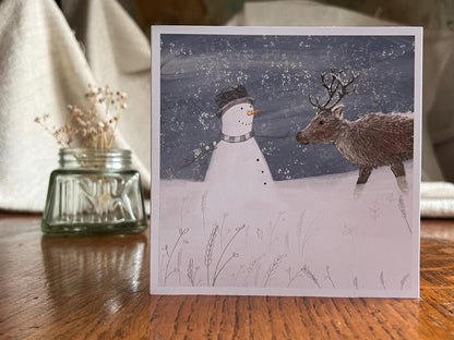 Premium illustrated Christmas greeting card featuring a happy snowman wearing a hat looking at a reindeer in a snowy winter landscape, displayed standing upright on a tabletop. Festive wildlife-inspired seasonal greeting card design.