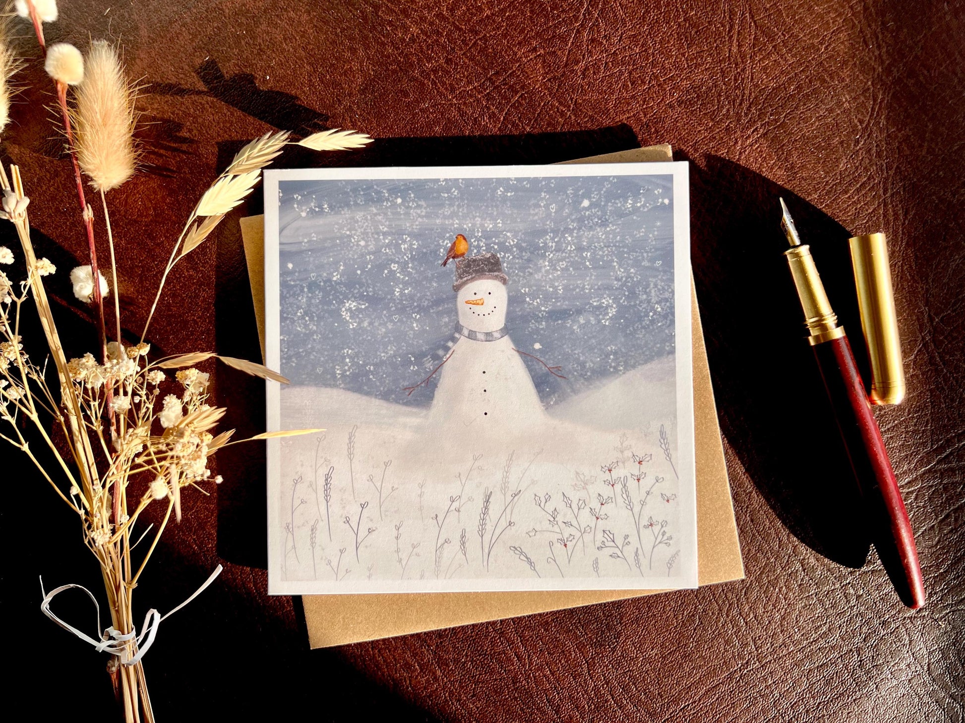  Premium illustrated Christmas greeting card featuring a happy snowman wearing a hat with a robin perched on top, set in a cheerful snowy winter landscape, styled in a flat lay on a leatherette tabletop with a recycled fleck kraft envelope, fountain pen, and dried flowers. Festive wildlife-inspired seasonal greeting card design.