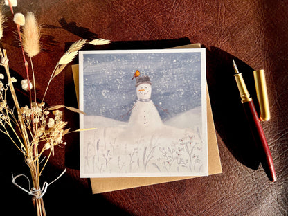  Premium illustrated Christmas greeting card featuring a happy snowman wearing a hat with a robin perched on top, set in a cheerful snowy winter landscape, styled in a flat lay on a leatherette tabletop with a recycled fleck kraft envelope, fountain pen, and dried flowers. Festive wildlife-inspired seasonal greeting card design.