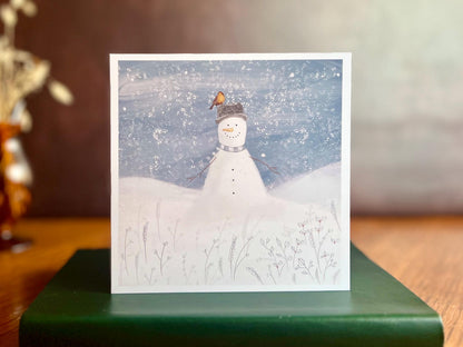 Premium illustrated Christmas greeting card featuring a happy snowman wearing a hat with a robin perched on top, set in a cheerful snowy winter landscape, displayed standing on a green leatherette book. Festive wildlife-inspired seasonal greeting card design.