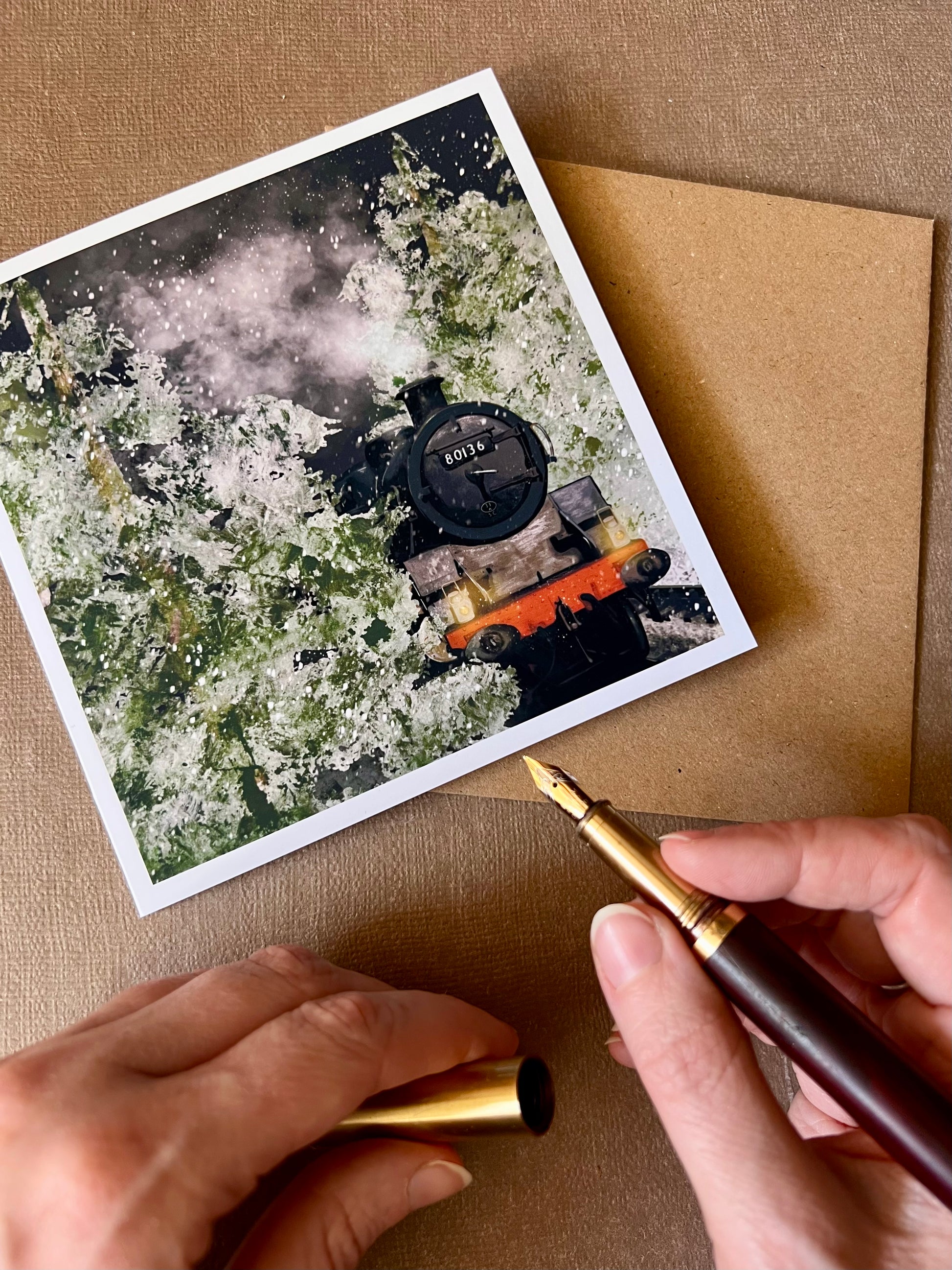 Premium illustrated Christmas greeting card featuring steam engine 80136 travelling through a snow-covered forest at night with frosted trees and steam rising into the night sky, shown on a tabletop with a hand holding a fountain pen about to write inside the card. Festive railway-themed winter scene card for train enthusiasts.