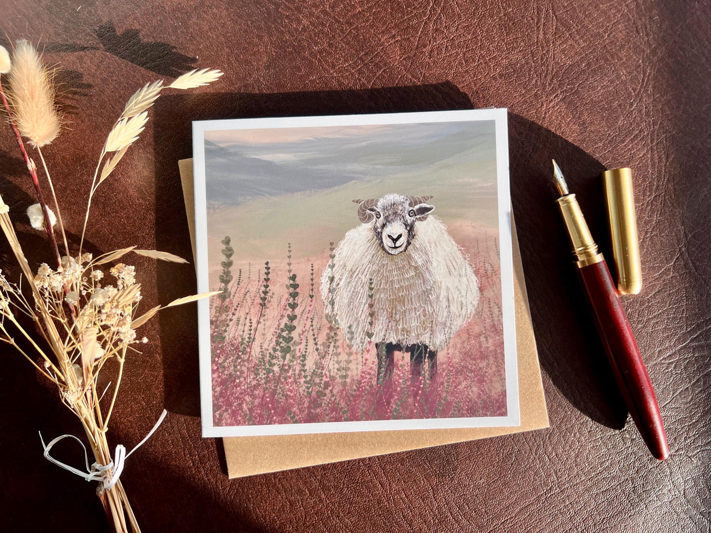 Premium illustrated art greeting card featuring a Swaledale sheep standing in a heather landscape on the Yorkshire moors, styled in a flat lay with dried flowers and a pen. Versatile countryside wildlife design with no text on the front, suitable for all occasions.