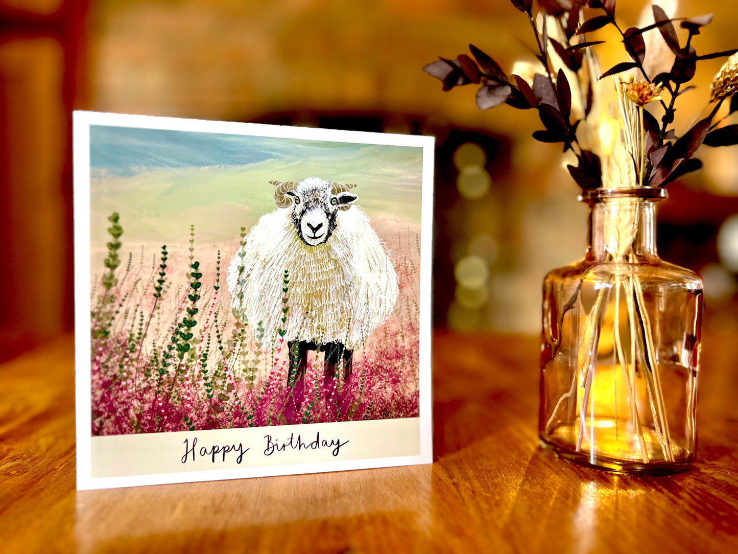 Premium illustrated birthday greeting card featuring a Swaledale sheep standing in a heather landscape on the Yorkshire moors, displayed standing upright on a rustic wooden tabletop. Countryside wildlife art card design with “Happy Birthday” text beneath the illustration.