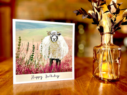 Premium illustrated birthday greeting card featuring a Swaledale sheep standing in a heather landscape on the Yorkshire moors, displayed standing upright on a rustic wooden tabletop. Countryside wildlife art card design with “Happy Birthday” text beneath the illustration.