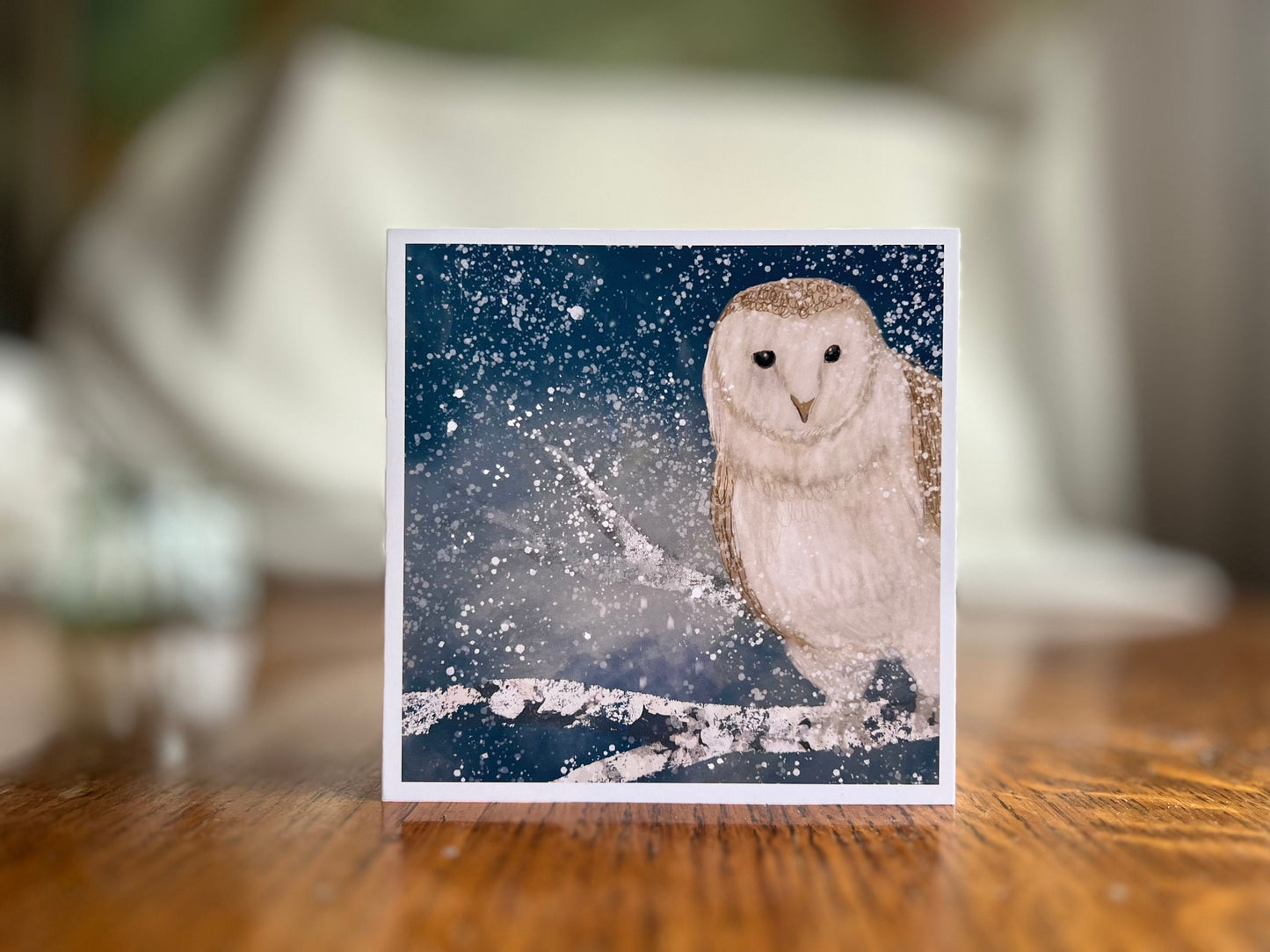 Premium high quality winter owl greeting card standing on a wooden tabletop with a softly blurred background, featuring an illustrated barn owl perched on a snowy branch under a magical blue and green night sky with falling snow, an ethereal artist-designed animal art card.