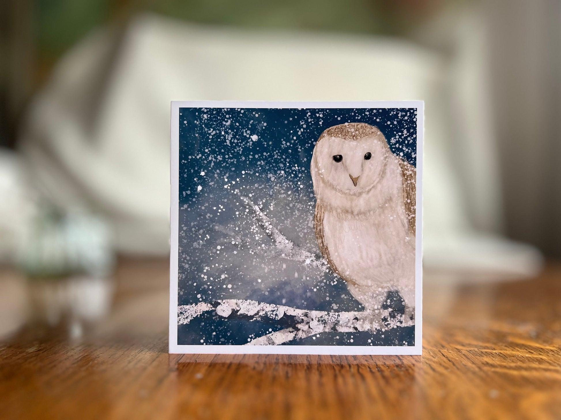 Premium high quality winter owl greeting card standing on a wooden tabletop with a softly blurred background, featuring an illustrated barn owl perched on a snowy branch under a magical blue and green night sky with falling snow, an ethereal artist-designed animal art card.