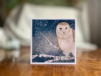 Premium high quality winter owl greeting card standing on a wooden tabletop with a softly blurred background, featuring an illustrated barn owl perched on a snowy branch under a magical blue and green night sky with falling snow, an ethereal artist-designed animal art card.