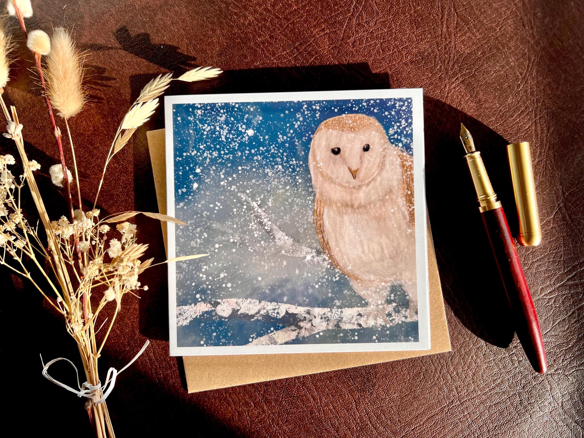 Premium high quality winter owl greeting card laid flat on a leatherette tabletop with a recycled kraft envelope underneath and a fountain pen to the right, with a bunch of dried flowers to the left, featuring an illustrated barn owl on a snowy branch under a magical blue and green winter night sky with falling snow, ethereal artist-designed animal art card.