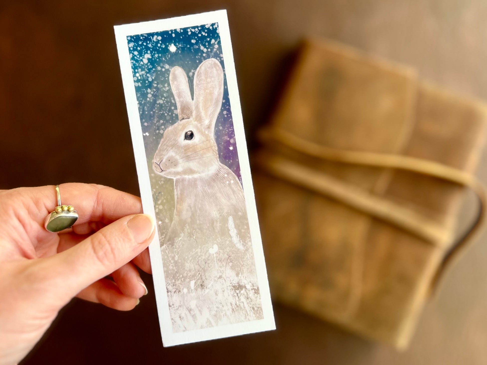 Hand holding a handcrafted rabbit bookmark featuring a celestial night sky illustration with a magical winter inspired atmosphere and soft glowing tones.