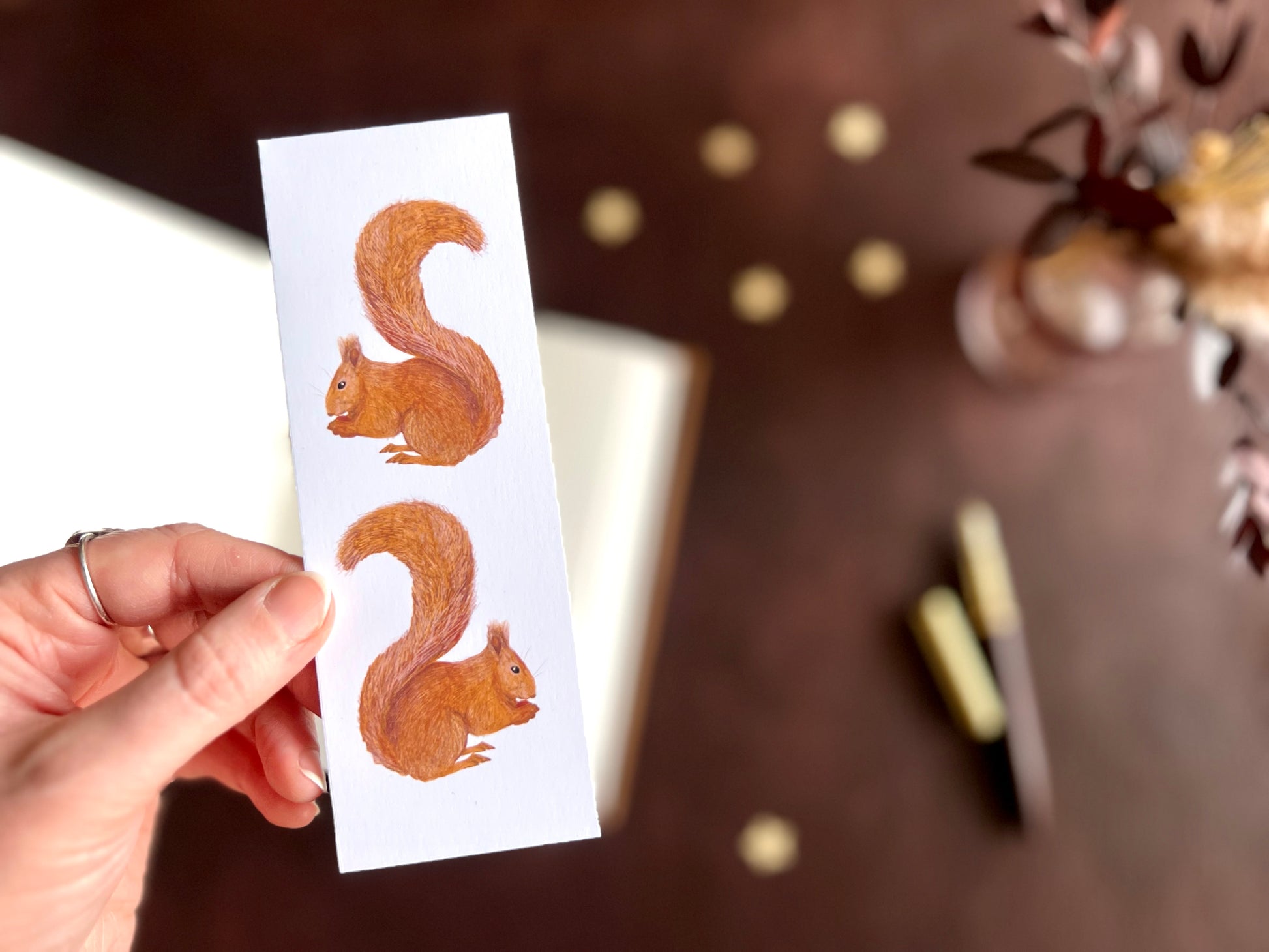 Hand holding a red squirrel bookmark styled with a fountain pen and open sketchbook in a cosy dark academia inspired desk scene.