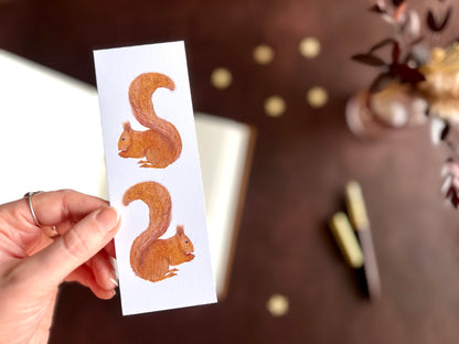 Hand holding a red squirrel bookmark styled with a fountain pen and open sketchbook in a cosy dark academia inspired desk scene.