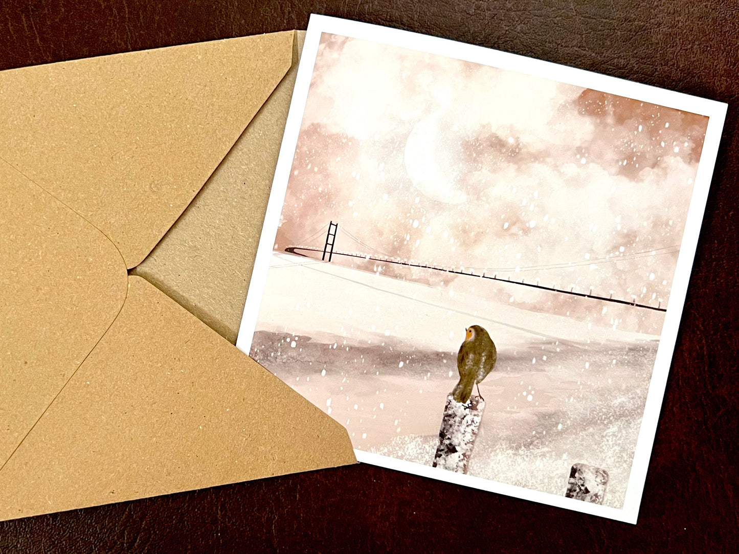 Close-up of a robin perched in a winter scene overlooking the River Humber, with the Humber Bridge softly visible in the background on a pink sepia toned greeting card.