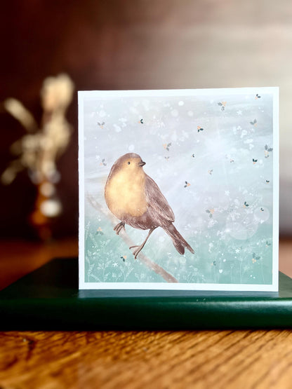 Premium winter greeting card featuring an illustrated robin perched on a branch against a soft wintry blue background with subtle holly leaf pattern and gentle sun rays through clouds. The card is shown standing on a book in a styled display. Ideal Christmas card or seasonal greeting for bird lovers and nature inspired festive gifting. Printed on thick 312gsm fine art card by a UK independent artist.