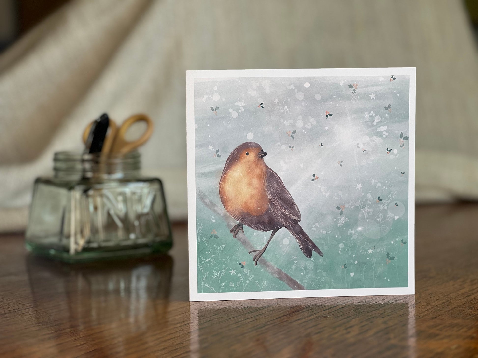 Premium winter greeting card featuring an illustrated robin perched on a branch against a soft wintry blue background with subtle holly leaf pattern and gentle sun rays. The card is shown standing on a rustic wooden tabletop. Ideal Christmas card or seasonal greeting for bird lovers and nature inspired festive gifting. Printed on thick 312gsm fine art card by a UK independent artist.