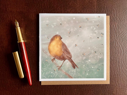 Premium winter greeting card featuring an illustrated robin perched on a branch against a soft wintry blue background with subtle holly leaf pattern and gentle sun rays through clouds. The card is shown laid flat on a tabletop. Ideal Christmas card or seasonal greeting for bird lovers and nature inspired festive gifting. Printed on thick 312gsm fine art card by a UK independent artist.