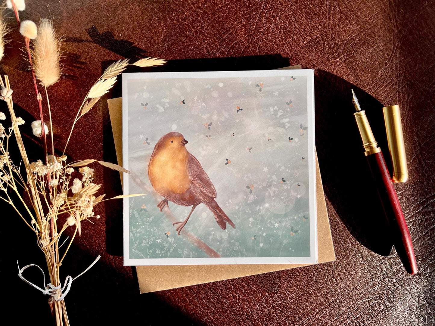 Premium winter greeting card featuring an illustrated robin perched on a branch against a soft wintry blue background with subtle holly leaf pattern and gentle sun rays. The card is shown laid flat with a pen and dried flowers on a tabletop in natural sunlight. Ideal Christmas card or seasonal greeting for bird lovers and nature inspired festive gifting. Printed on thick 312gsm fine art card by a UK independent artist.