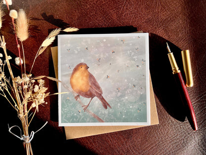 Premium winter greeting card featuring an illustrated robin perched on a branch against a soft wintry blue background with subtle holly leaf pattern and gentle sun rays. The card is shown laid flat with a pen and dried flowers on a tabletop in natural sunlight. Ideal Christmas card or seasonal greeting for bird lovers and nature inspired festive gifting. Printed on thick 312gsm fine art card by a UK independent artist.