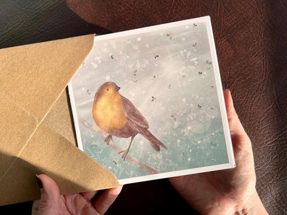 Premium winter greeting card featuring an illustrated robin perched on a branch against a soft wintry blue background. The design includes a subtle holly leaf pattern and gentle sun rays breaking through winter clouds. Ideal as a Christmas card or seasonal greeting for bird lovers and nature inspired festive gifting. Printed on thick 312gsm fine art card by a UK independent artist.