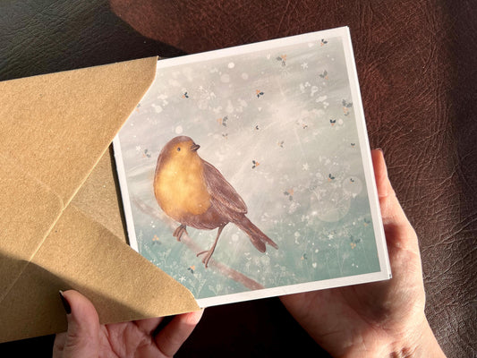 Premium winter greeting card featuring an illustrated robin perched on a branch against a soft wintry blue background. The design includes a subtle holly leaf pattern and gentle sun rays breaking through winter clouds. Ideal as a Christmas card or seasonal greeting for bird lovers and nature inspired festive gifting. Printed on thick 312gsm fine art card by a UK independent artist.
