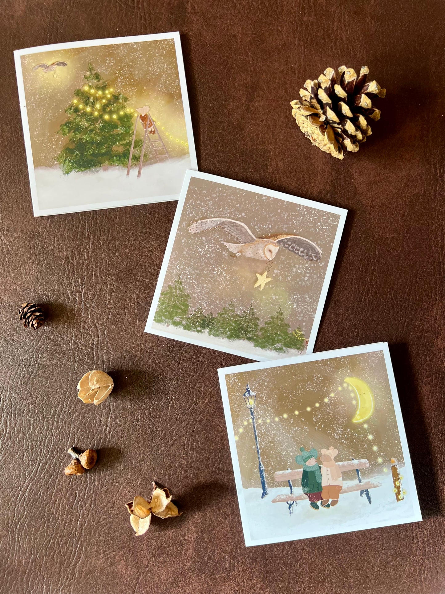 Premium set of three Christmas cards featuring storybook-style woodland winter illustrations. Designs include mice in winter coats on a snowy bench, a barn owl carrying a smiling star through a snowy sky, and a mouse decorating a Christmas tree with fairy lights. The cards are shown laid flat on a brown leatherette surface with pine cones and woodland decorations. Ideal Christmas card set for animal lovers and rustic festive gifting. Printed on thick 312gsm fine art card by a UK independent artist.
