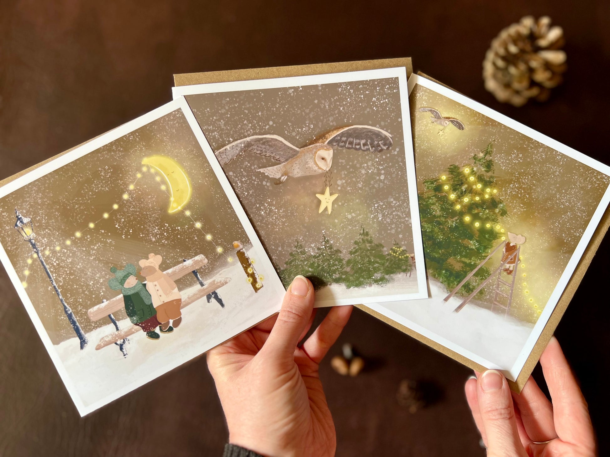 Premium set of three Christmas cards featuring storybook-style woodland winter illustrations. Designs include two mice in winter hats sitting on a bench beneath a fairy-lit moon, a barn owl carrying a smiling star through a snowy sky, and a mouse decorating a Christmas tree with fairy lights. The cards are shown in a fan shape being held in two hands to display all three designs. Ideal Christmas card pack for animal lovers and rustic winter gifting. Printed on thick 312gsm fine art card by a UK artist