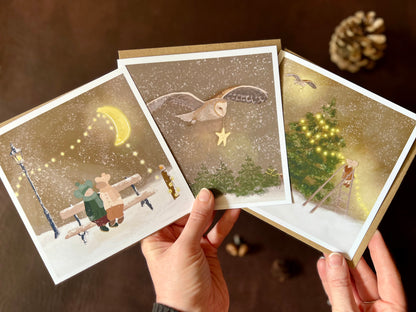 Premium set of three Christmas cards featuring storybook-style woodland winter illustrations. Designs include two mice in winter hats sitting on a bench beneath a fairy-lit moon, a barn owl carrying a smiling star through a snowy sky, and a mouse decorating a Christmas tree with fairy lights. The cards are shown in a fan shape being held in two hands to display all three designs. Ideal Christmas card pack for animal lovers and rustic winter gifting. Printed on thick 312gsm fine art card by a UK artist
