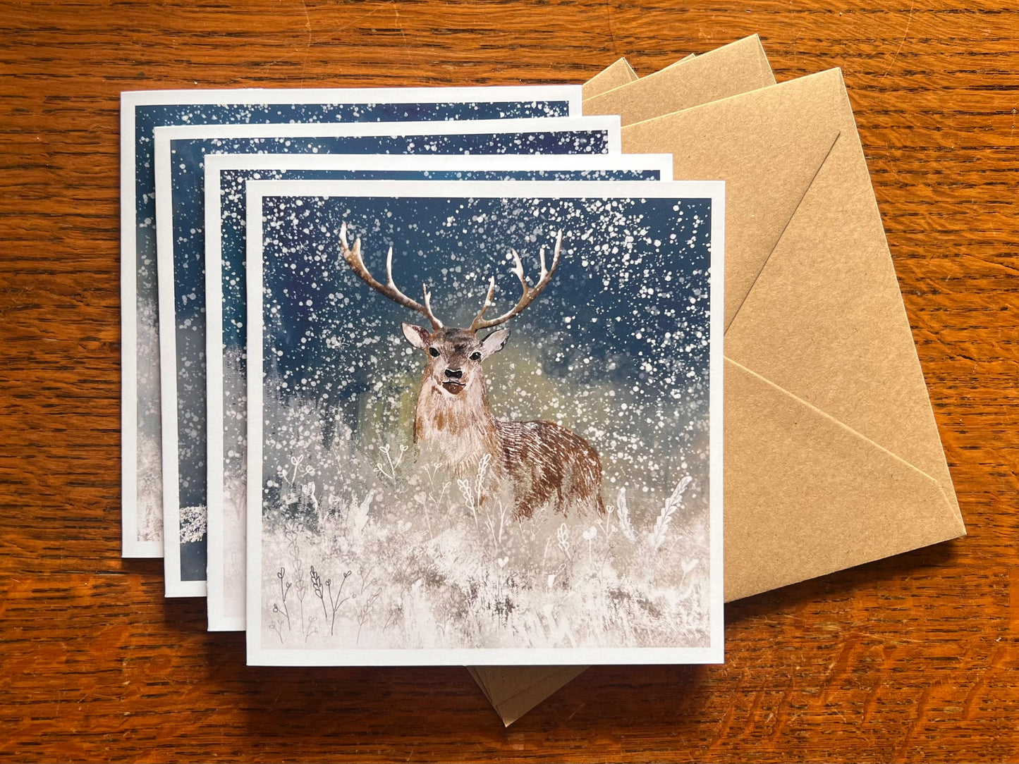 Set of four winter animal greeting cards stacked on top of recycled kraft envelopes on a wooden tabletop, with the top card featuring an illustrated stag standing in snowy flowers under a blue and green winter night sky with falling snow, a magical ethereal Christmas and animal art greeting card set.