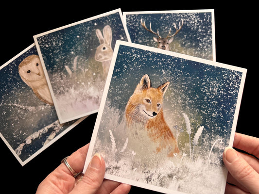 Hands holding a winter animal greeting card from a set of 4 cards, featuring an illustrated orange fox sitting among snowy flowers on a snowy winter’s night with an ethereal magical style, with three additional cards laid flat showing owl, rabbit and stag designs in the background, versatile Christmas or animal art greeting card set.