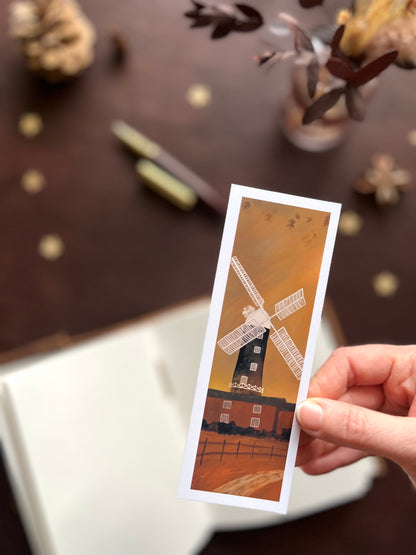 Close-up of a handcrafted Skidby Windmill bookmark featuring a countryside landscape with burnt orange sunset sky, photographed with soft blurred pine cones and flowers in the background.