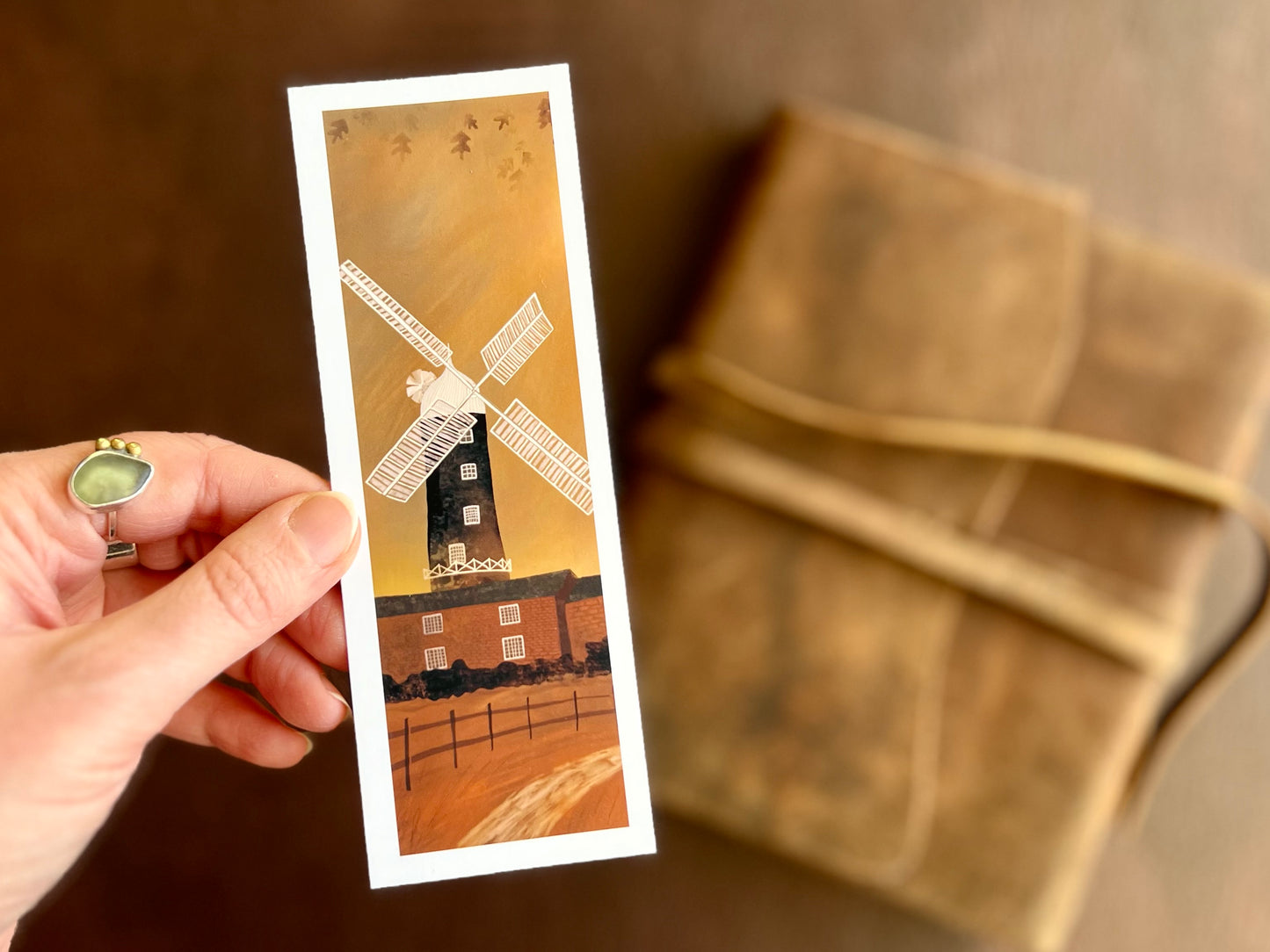 Hand holding a handcrafted Skidby Windmill bookmark featuring a countryside landscape with burnt orange autumn sunset sky and East Yorkshire windmill illustration.
