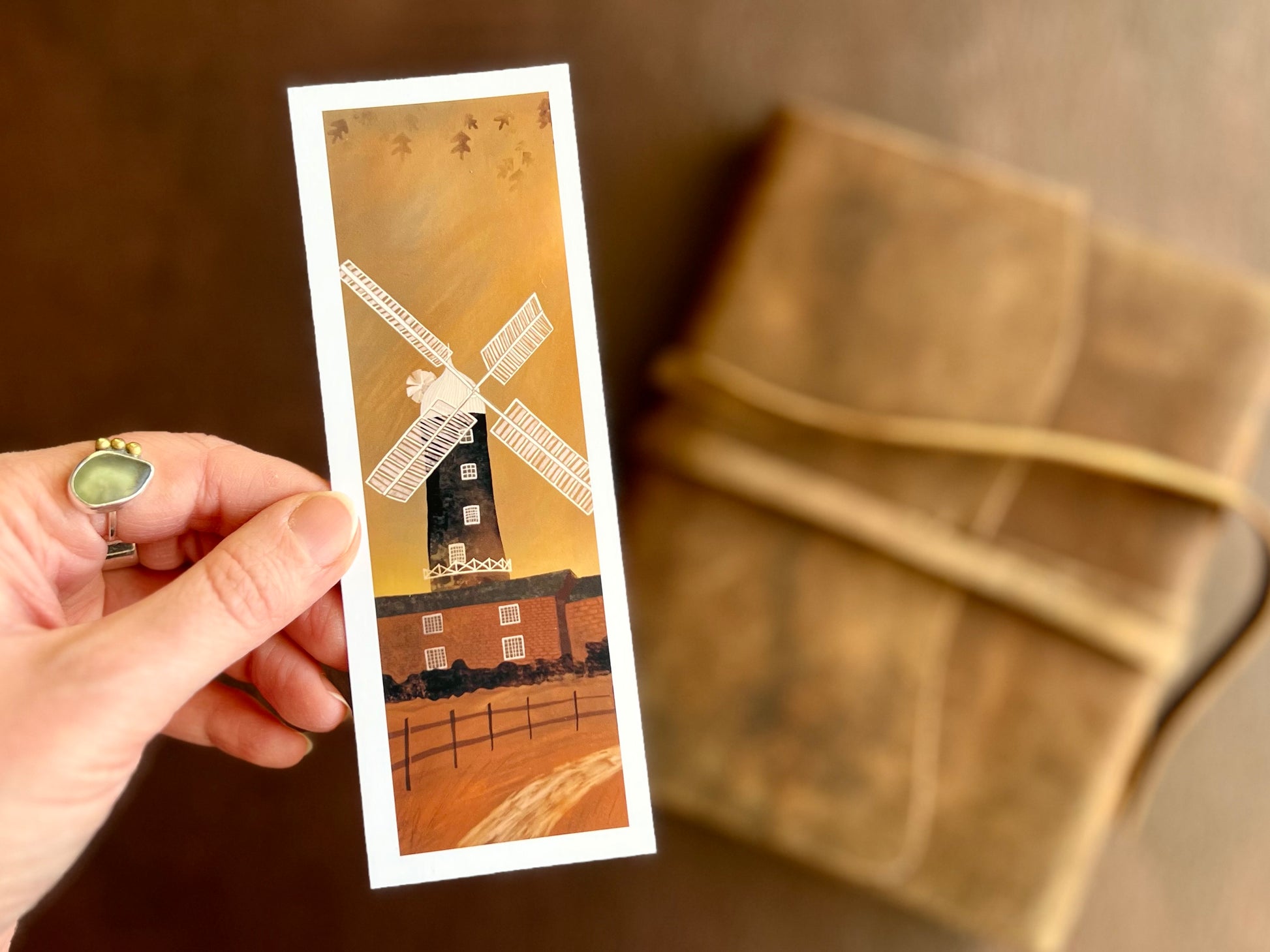 Hand holding a handcrafted Skidby Windmill bookmark featuring a countryside landscape with burnt orange autumn sunset sky and East Yorkshire windmill illustration.