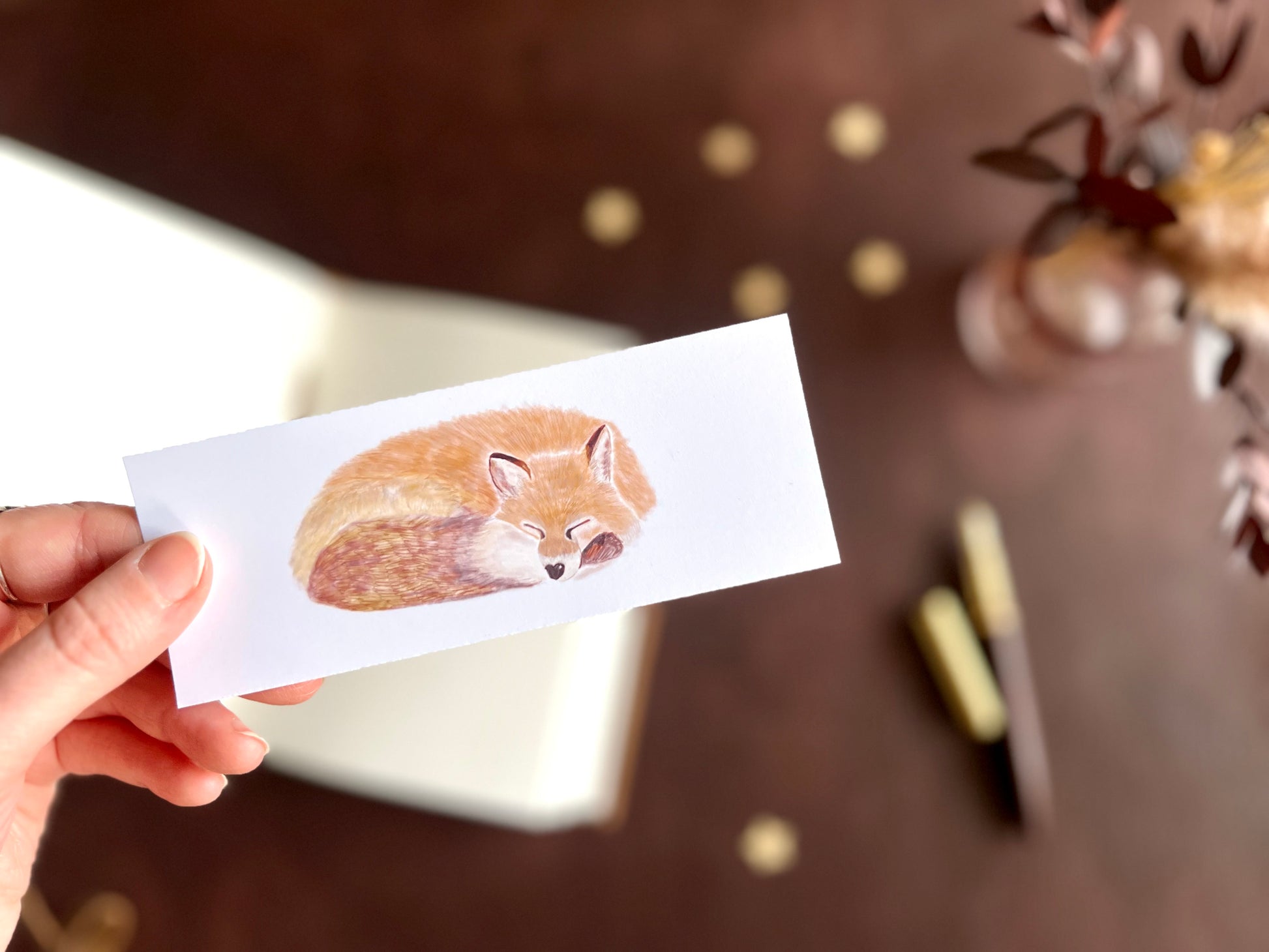 Sleeping fox bookmark shown with a fountain pen and open sketchbook on a cosy dark academia inspired writing desk.