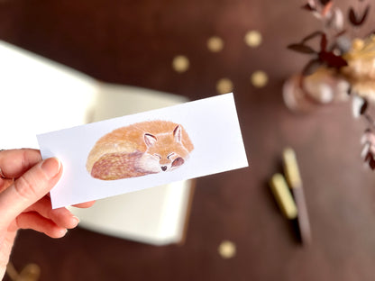 Sleeping fox bookmark shown with a fountain pen and open sketchbook on a cosy dark academia inspired writing desk.