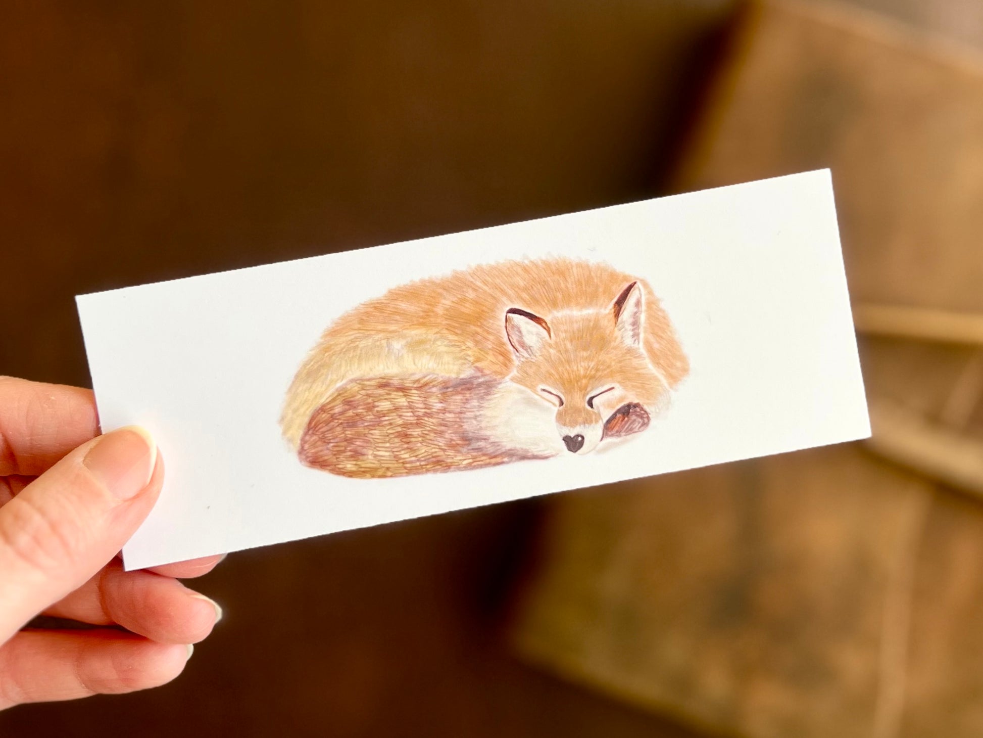 Hand holding a handcrafted sleeping fox bookmark featuring a curled-up fox illustration on a clean white background.