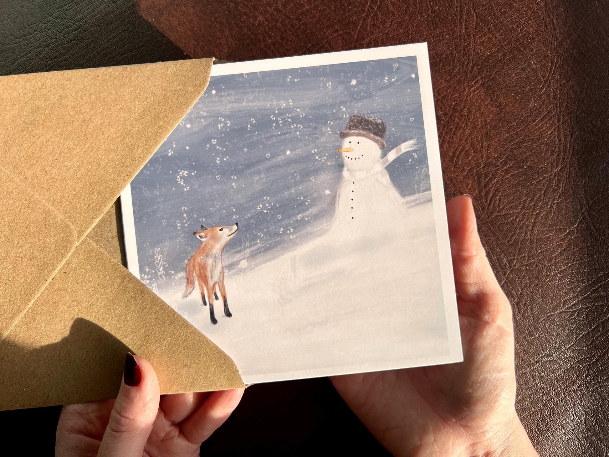 Premium Christmas card featuring a cheerful snowman wearing a hat sitting in a blustery snowy scene beside a small fox. The illustration has a storybook-style winter woodland feel. The card has no text and is shown in close-up being held in two hands. Ideal festive greeting card for animal lovers and seasonal winter gifting. Printed on thick 312gsm fine art card by a UK independent artist.