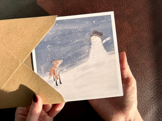 Premium Christmas card featuring a cheerful snowman wearing a hat sitting in a blustery snowy scene beside a small fox. The illustration has a storybook-style winter woodland feel. The card has no text and is shown in close-up being held in two hands. Ideal festive greeting card for animal lovers and seasonal winter gifting. Printed on thick 312gsm fine art card by a UK independent artist.