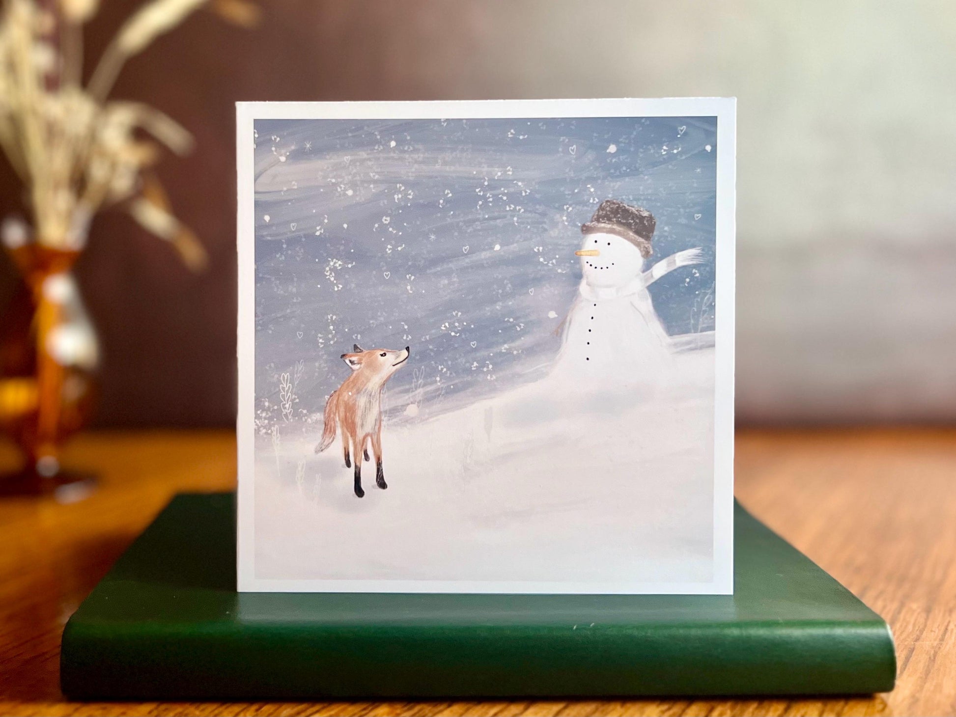 Premium Christmas card featuring a cheerful snowman wearing a hat sitting beside a small fox in a snowy winter scene. The background shows a blue snowy sky with a blustery seasonal feel. The card has no text and is shown standing on a book in a styled display. Ideal festive greeting card for animal lovers and winter gifting. Printed on thick 312gsm fine art card by a UK independent artist.
