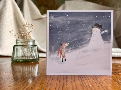 Premium Christmas card featuring a cheerful snowman wearing a hat sitting beside a small fox in a snowy winter scene with a blue snowy sky. The card has no text and is shown standing on a rustic wooden tabletop. Ideal festive greeting card for animal lovers and winter gifting. Printed on thick 312gsm fine art card by a UK independent artist.