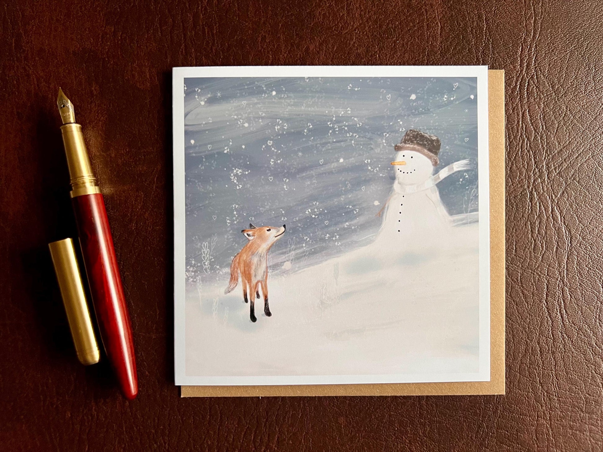 Premium Christmas card featuring a cheerful snowman wearing a hat sitting beside a small fox in a snowy winter scene with a blue snowy sky. The card has no text and is shown laid flat on a tabletop with a pen, highlighting space for a personal message inside. Ideal festive greeting card for animal lovers and winter gifting. Printed on thick 312gsm fine art card by a UK independent artist.