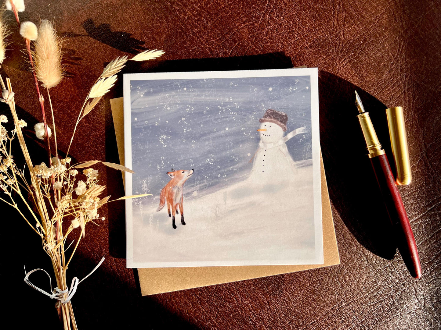 Premium Christmas card featuring a cheerful snowman wearing a hat sitting beside a small fox in a snowy winter scene with a blue snowy sky. The card has no text and is shown laid flat on a tabletop with a pen and dried flowers, highlighting space for a personal message inside. Ideal festive greeting card for animal lovers and winter gifting. Printed on thick 312gsm fine art card by a UK independent artist.