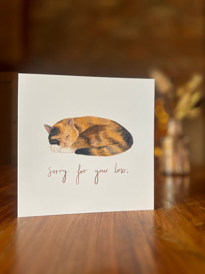 “Sorry for your loss” calico cat sympathy card featuring a sleeping tortoiseshell and white cat illustration, shown on a rustic wooden tabletop with softly blurred dried flowers and a brick fireplace. A special cat bereavement card, pet sympathy card, and condolence card for loss of a cat.