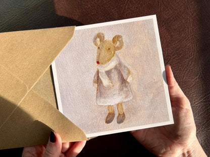 Hands holding an artisan greeting card being removed from a kraft envelope, featuring a print of an oil painting titled 'Mouse in a Lilac Dress'. The artwork shows a small brown mouse wearing a lilac dress with a white scarf and mittens against a matching lilac background.