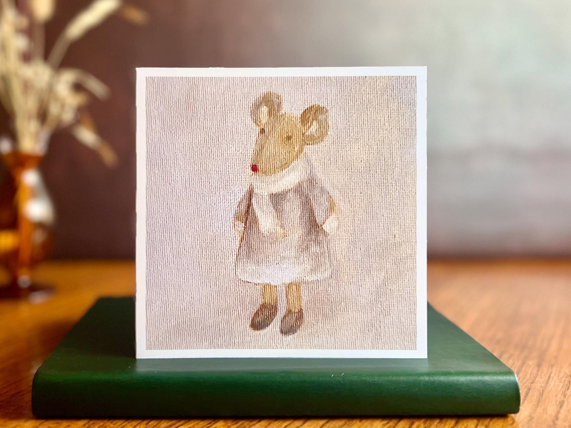 Artisan greeting card featuring a print of an oil painting titled 'Mouse in a Lilac Dress', showing a small brown mouse wearing a lilac dress with a white scarf and mittens, displayed standing on a green leatherette book in a styled lifestyle setting.